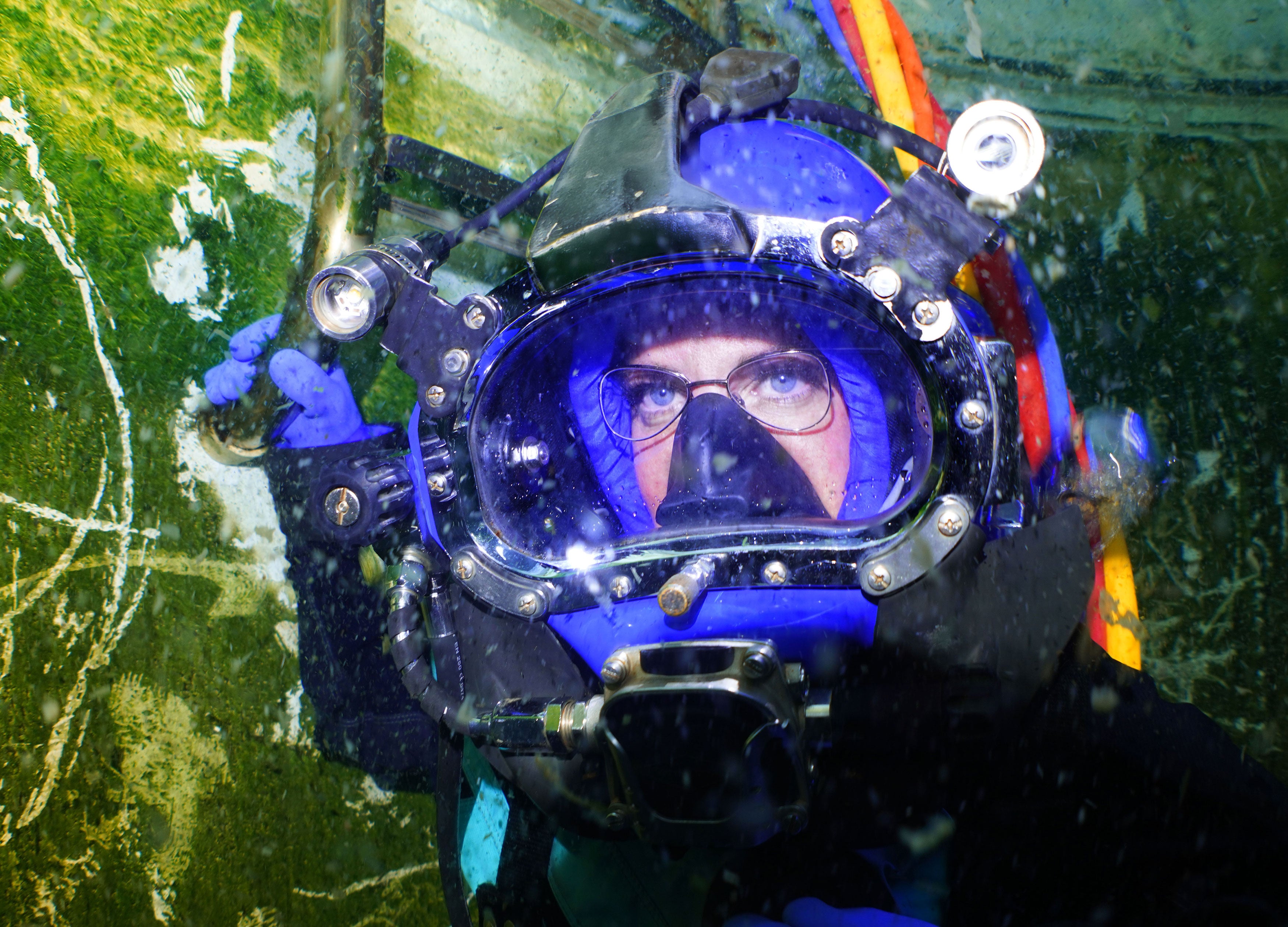 Person wearing a large diving helmet underwater with greenery in the background
