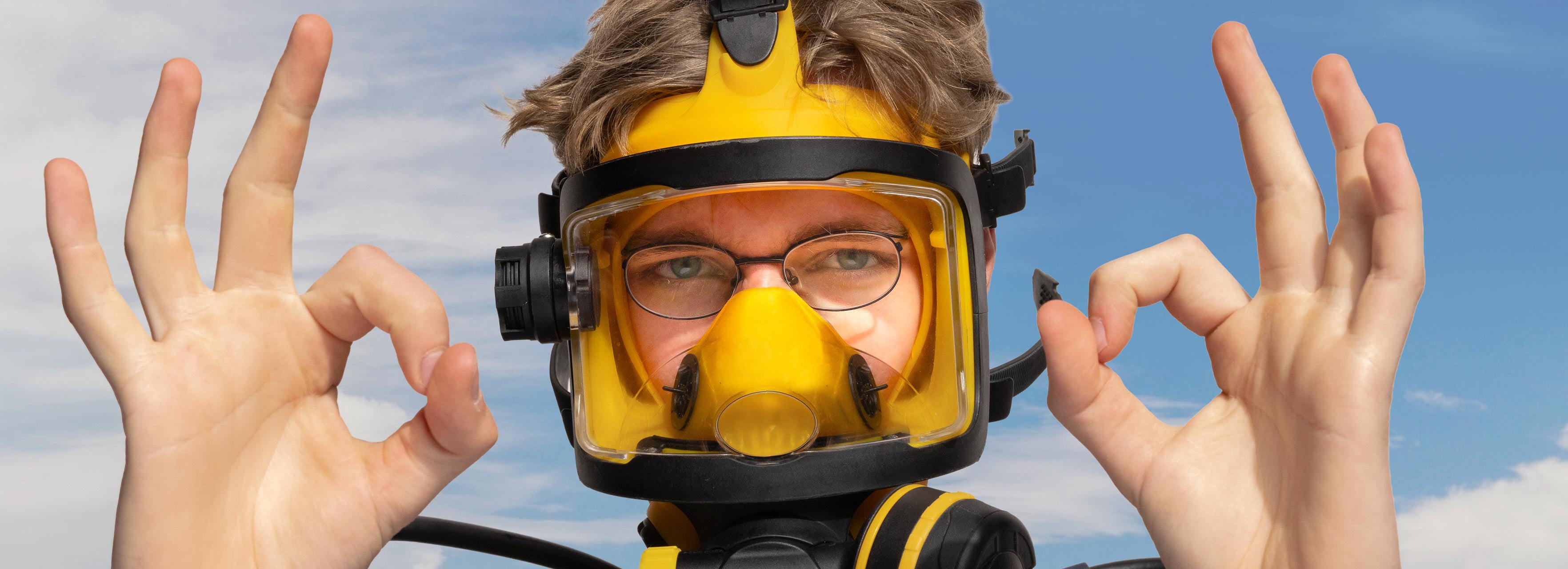 Person wearing a yellow diving mask with clear visor, making hand gestures against a blue sky.
